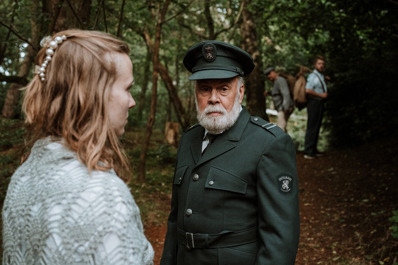 Douanebeambte in groen uniform spreekt vrouw aan in Brabants bos, met twee mannen op achtergrond.