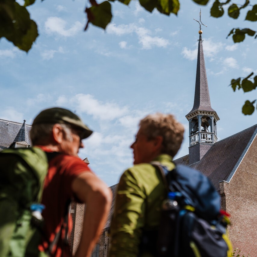 Twee wandelaars met rugzakken kijken naar een kerktoren in een Brabants dorp onder een blauwe lucht.