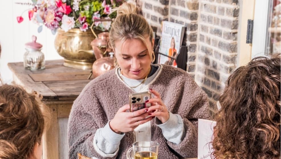 Drie vrouwen drinken thee en eten taart in een knusse Brabantse lunchroom met bakstenen muur en bloemen.