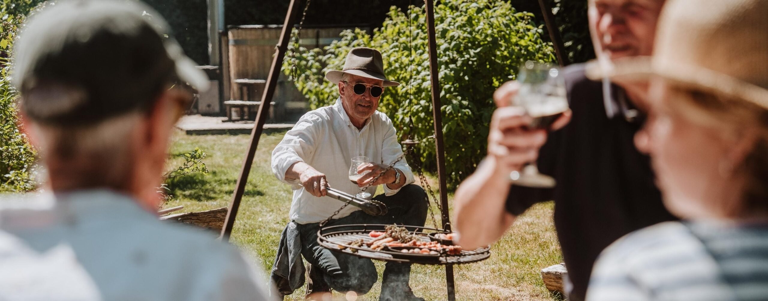 Man grilt groenten op een buitenbarbecue terwijl gasten genieten van drankjes in een zonnige tuin in Brabant.