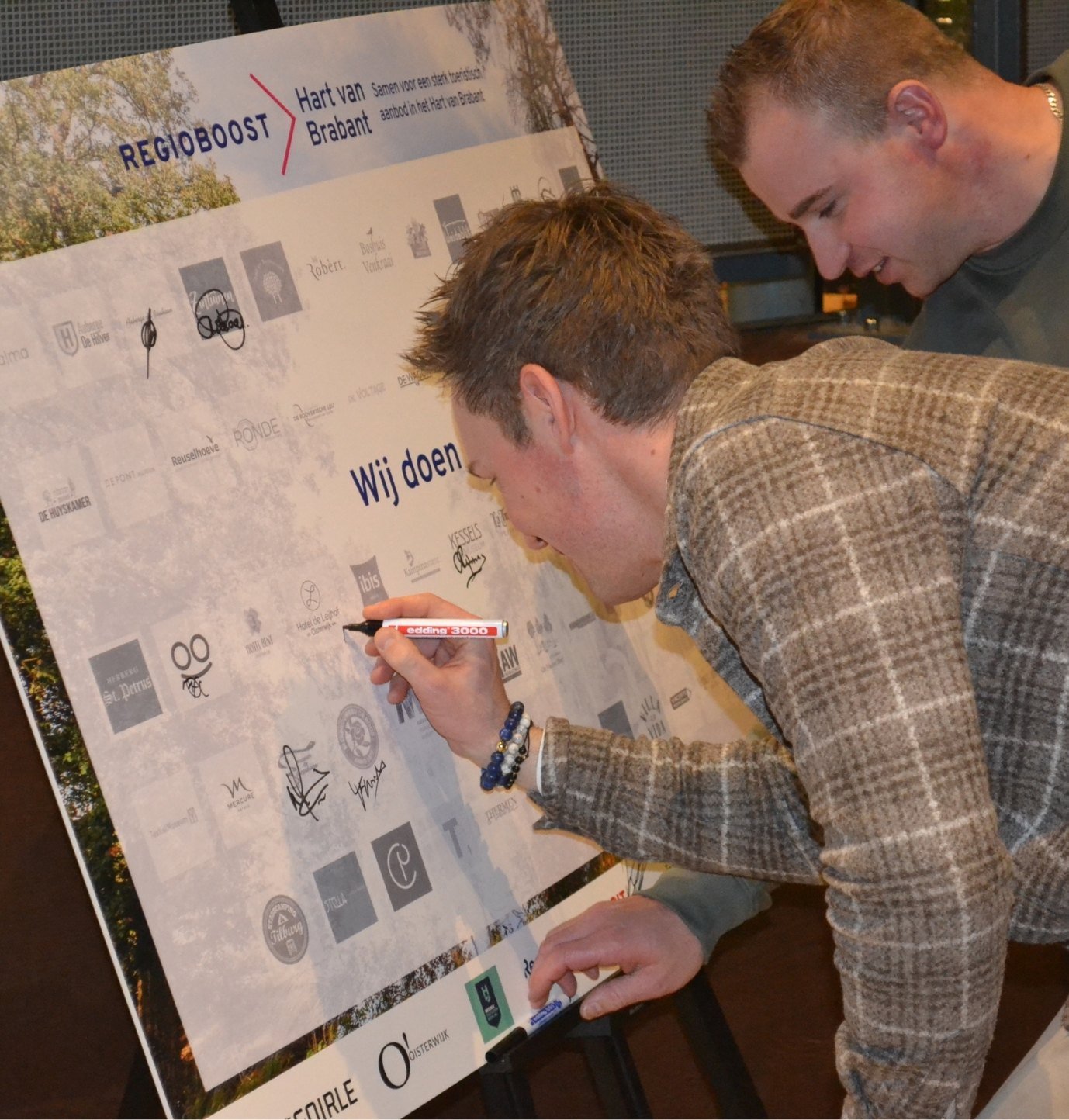 Twee mannen zetten een handtekening op het RegioBoost-bord van Hart van Brabant tijdens een samenwerkingsmoment.