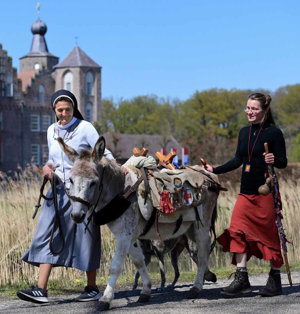 Twee vrouwen wandelen met een beladen ezel langs Kasteel Heeswijk in Brabant op een zonnige dag.