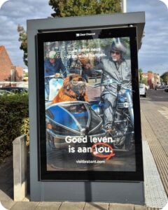 Abri met campagneposter van Goed Leven van Brabant Partners met man en hond in zijspan op motorrit door een Brabants dorp.